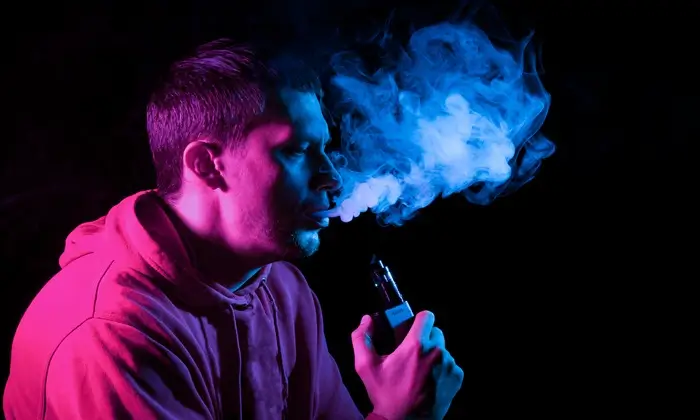 close up portrait face adult serious man exhales blue toxic smoke while smoking ecigarette vape illuminated with pink colored light black background harm health
